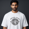 A man wearing a white crew-neck t-shirt with a black graphic print that reads 'JUGAAD EXPERT' around a circular maze design, set against a dark gray studio background.