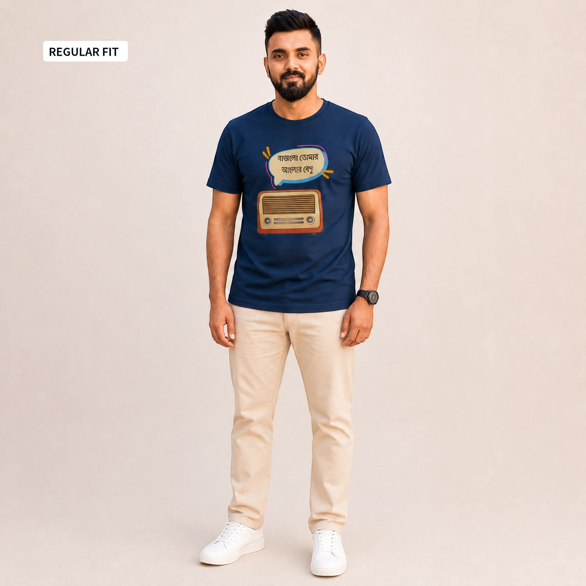 A man modeling a navy blue graphic t-shirt with a retro radio and Bengali text design, paired with beige trousers and white sneakers, against a solid light beige background. A 'REGULAR FIT' label is visible in the top-left corner.