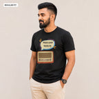 A man wearing a black short-sleeve t-shirt with a graphic print of a retro radio and Bengali text inside a speech bubble, paired with beige trousers, standing against a plain light beige background.