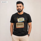 A man wearing a black graphic t-shirt with a retro radio and Bengali text design, paired with beige trousers, standing against a plain beige background.