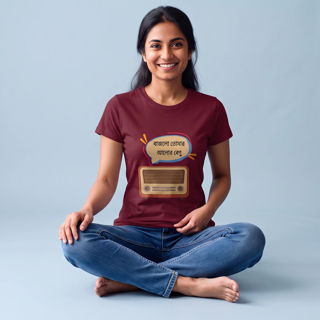 A woman sitting cross-legged on a light blue studio background wearing a maroon graphic t-shirt with a cartoon radio and Bengali text speech bubble, paired with blue jeans.