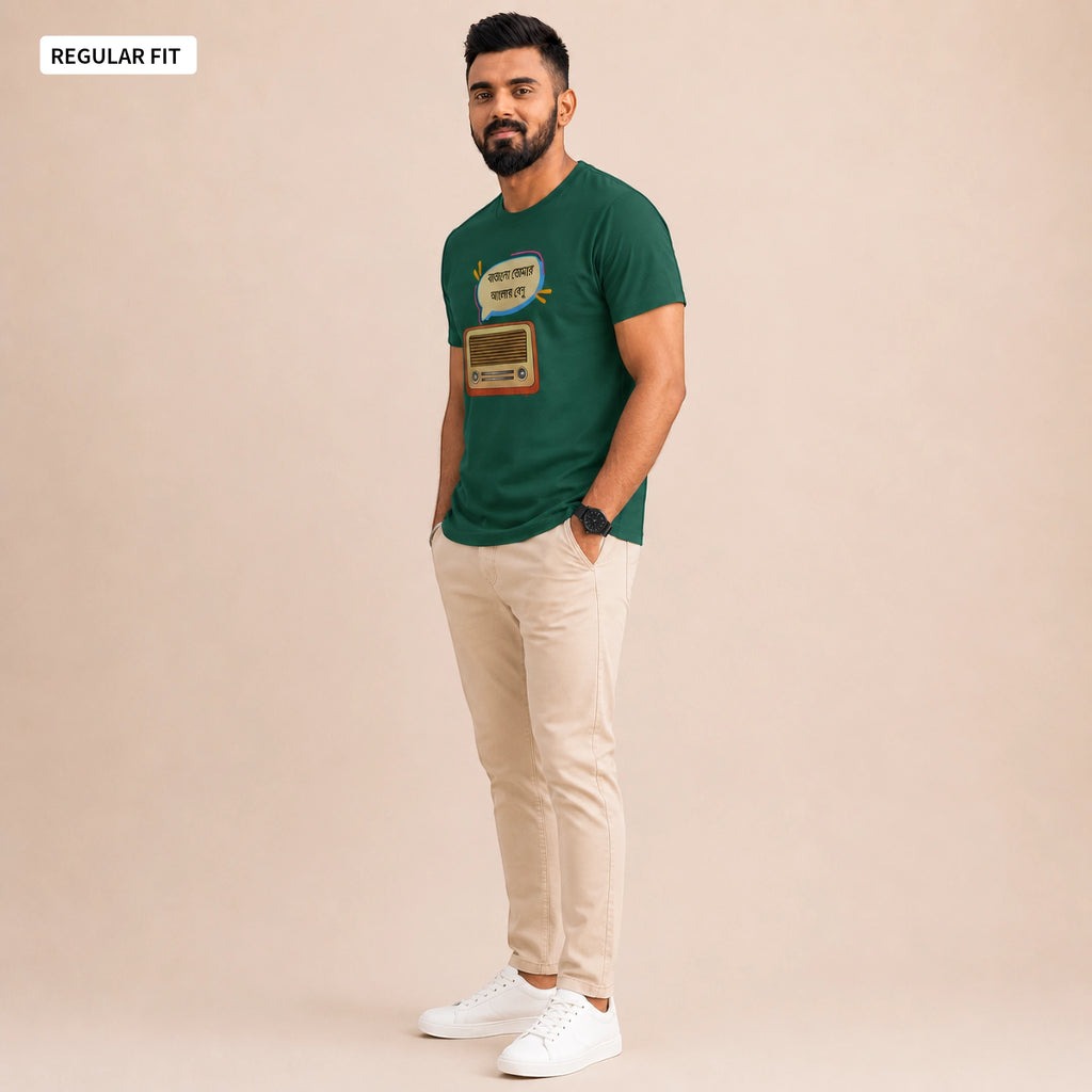 A man modeling a dark green graphic t-shirt with a retro radio design, paired with beige trousers and white sneakers, set against a solid beige studio background.