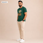 A man modeling a dark green graphic t-shirt with a retro radio design, paired with beige trousers and white sneakers, set against a solid beige studio background.