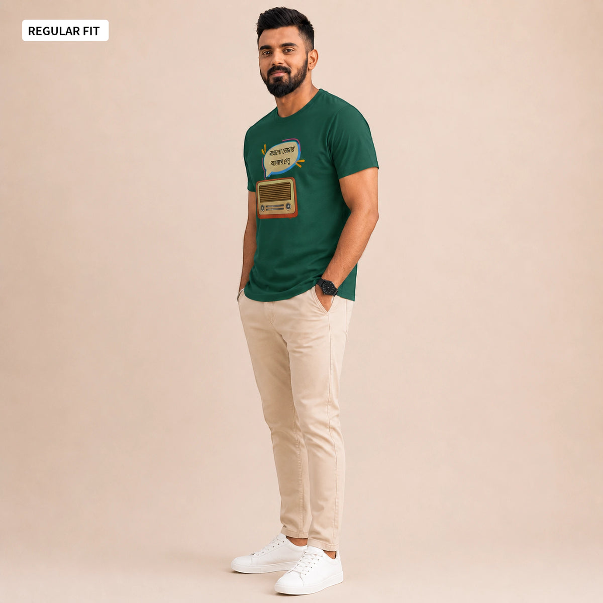 A man modeling a dark green graphic t-shirt with a retro radio design, paired with beige trousers and white sneakers, set against a solid beige studio background.