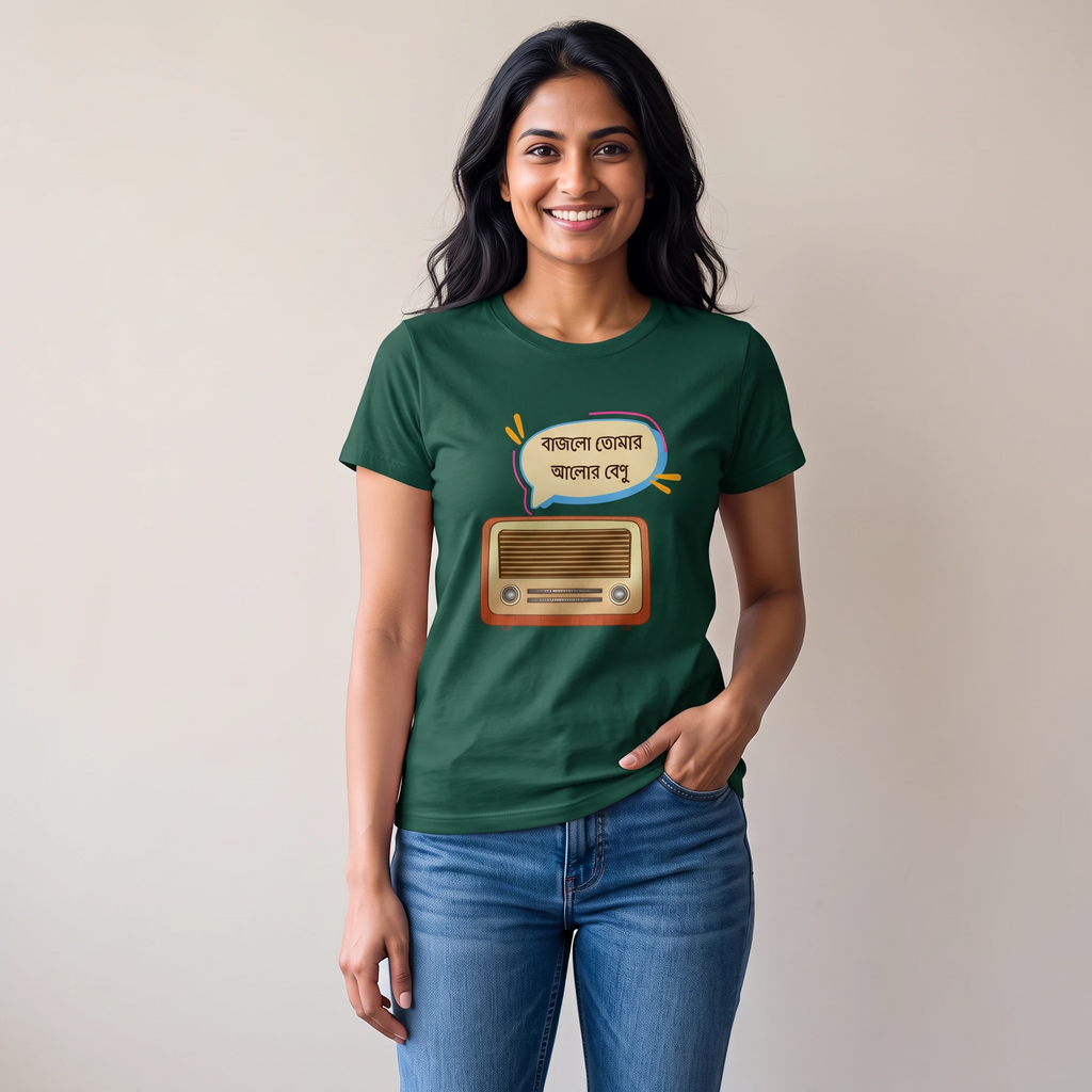 A woman wearing a dark green graphic t-shirt with a cartoon radio and Bengali text print, paired with blue jeans, standing against a plain beige wall.