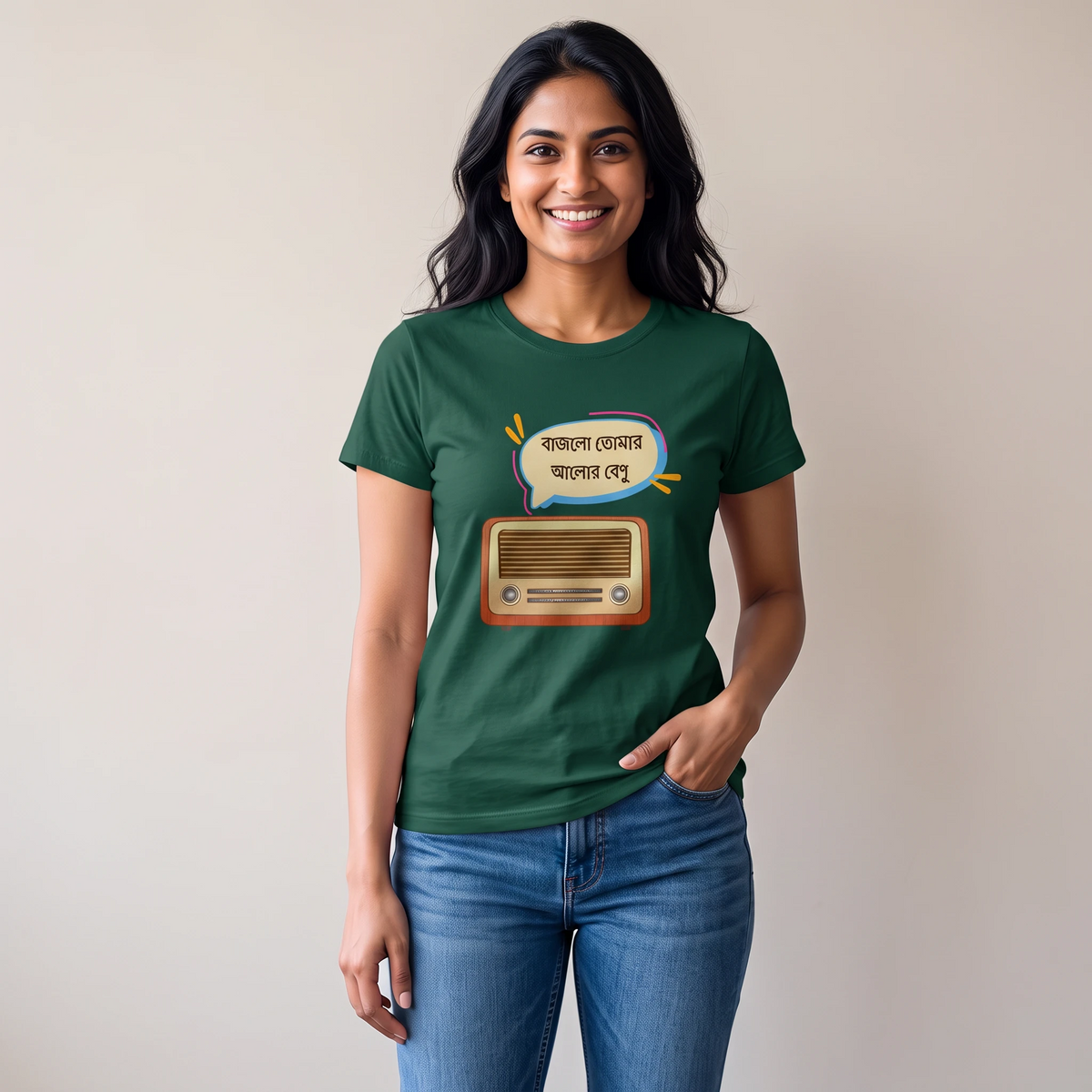 A woman wearing a dark green graphic t-shirt with a cartoon radio and Bengali text print, paired with blue jeans, standing against a plain beige wall.
