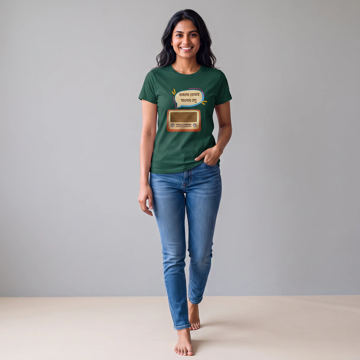 A woman stands barefoot against a light gray studio background wearing a dark green graphic T-shirt and blue jeans. The T-shirt has a retro television graphic with a speech bubble that reads 'bajlo tomar alor benu'