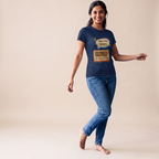 A woman modeling a navy blue graphic t-shirt with a retro radio design and the text 'Bajlo tomar alor benu'. She is wearing blue jeans and standing barefoot against a solid beige studio background.