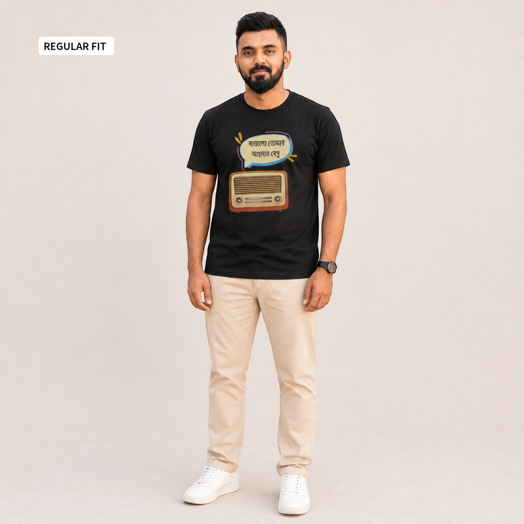 A man modeling a black graphic t-shirt with a retro radio design and Bengali text, paired with beige trousers and white sneakers, against a light beige background. A 'REGULAR FIT' label is visible in the top-left corner.