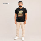 A man modeling a black graphic t-shirt with a retro radio design and Bengali text, paired with beige trousers and white sneakers, against a light beige background. A 'REGULAR FIT' label is visible in the top-left corner.