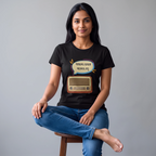 A woman sitting on a wooden stool wearing a black graphic t-shirt with a retro radio and speech bubble design in Bengali, paired with blue jeans, against a solid gray studio background.