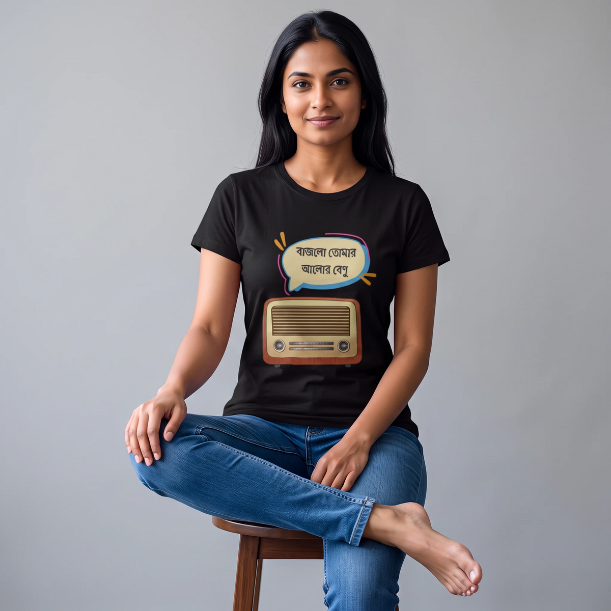 A woman sitting on a wooden stool wearing a black graphic t-shirt with a retro radio and speech bubble design in Bengali, paired with blue jeans, against a solid gray studio background.