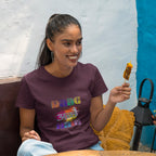 Woman wearing a purple t-shirt with colorful text, holding a skewer, sitting against a blue wall.