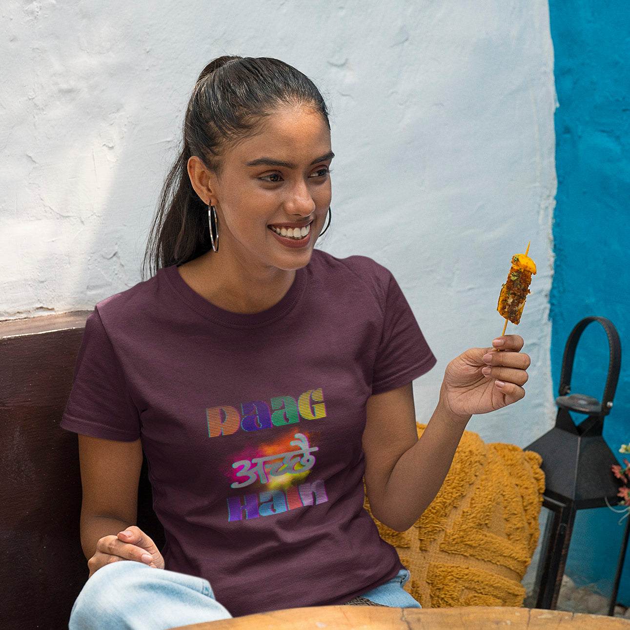 Woman wearing a purple t-shirt with colorful text, holding a skewer, sitting against a blue wall.