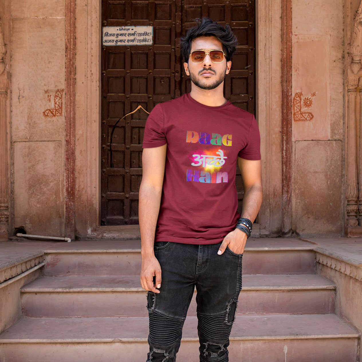 Man wearing a maroon t-shirt with colorful text standing on steps in front of an old building.