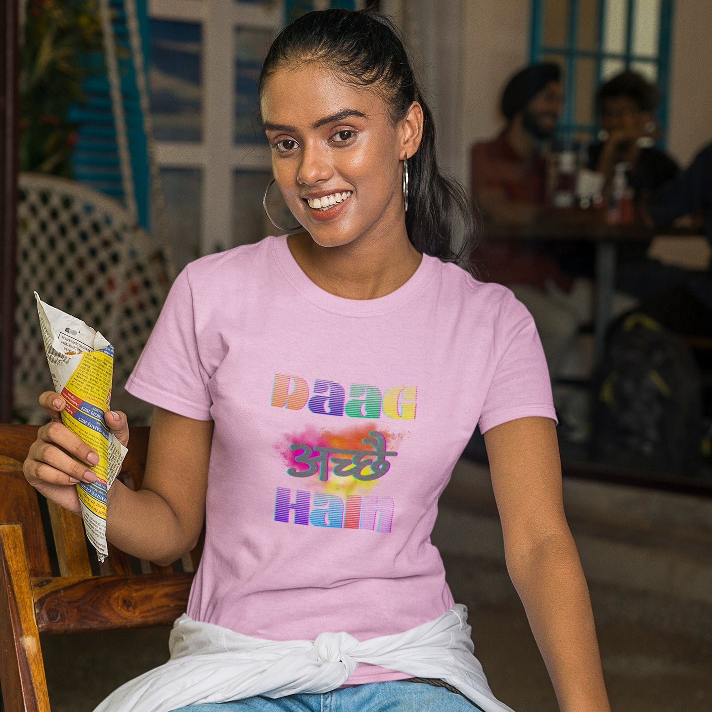Woman wearing a pink t-shirt with colorful text, holding a snack packet indoors.