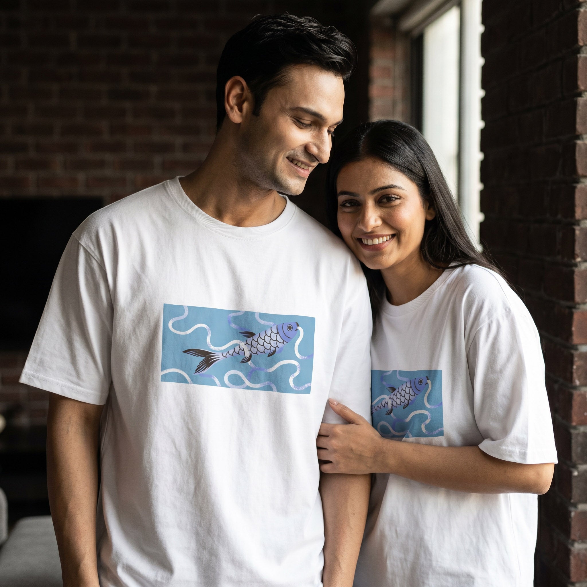 A man and a woman standing indoors wearing matching white T-shirts with a blue rectangular graphic of two fish on the front.