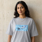 A woman wearing a light gray oversized t-shirt with a graphic print of a blue and white fish swimming in wavy water, standing against a textured concrete wall.