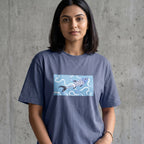 A woman wearing a slate blue crewneck t-shirt with a graphic print of a koi fish swimming in a light blue rectangular box on the chest, set against a textured gray concrete wall.
