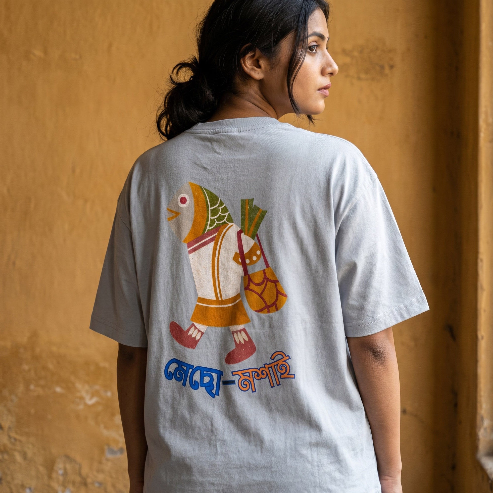 A woman shown from the back wearing a light gray t-shirt with a colorful graphic of a cartoon fish going to the market, with the words 'Mecho Moshai' written below in blue and orange. She is standing against a textured yellow wall.