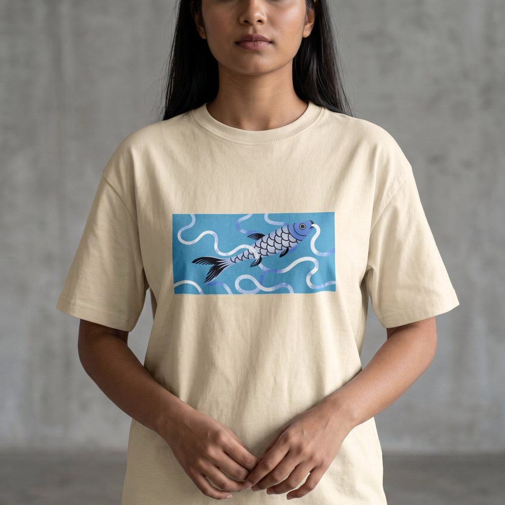 A woman wearing a cream-colored t-shirt with a graphic of a blue and purple koi fish on a light blue background, paired with blue jeans, standing against a gray concrete wall.