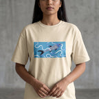 A woman wearing a cream-colored t-shirt with a graphic of a blue and purple koi fish on a light blue background, paired with blue jeans, standing against a gray concrete wall.