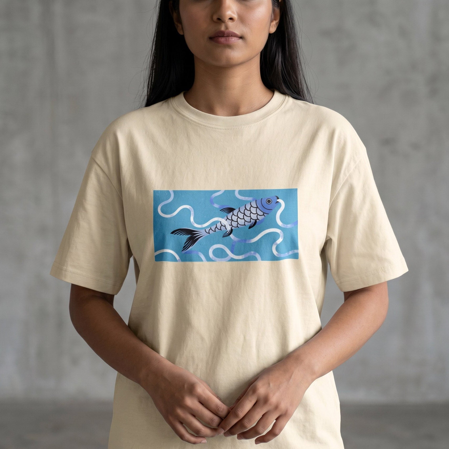 A woman wearing a cream-colored t-shirt with a graphic of a blue and purple koi fish on a light blue background, paired with blue jeans, standing against a gray concrete wall.