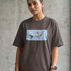 A woman wearing an oversized dark brown t-shirt with a rectangular graphic print of a koi fish on a light blue background, standing against a textured concrete wall.