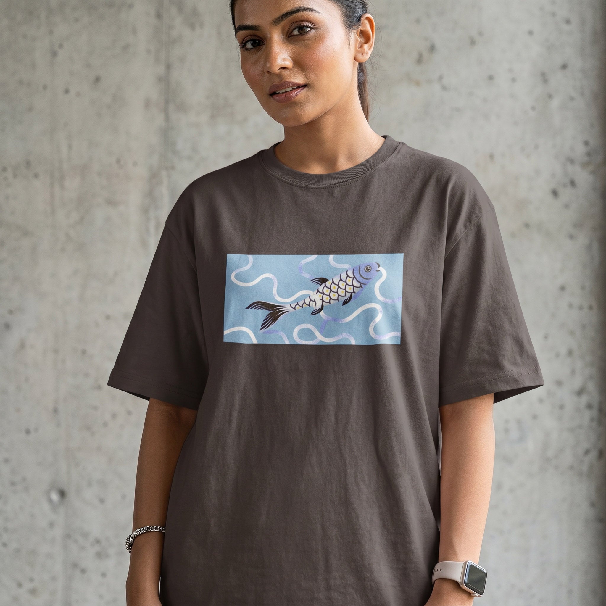 A woman wearing an oversized dark brown t-shirt with a rectangular graphic print of a koi fish on a light blue background, standing against a textured concrete wall.