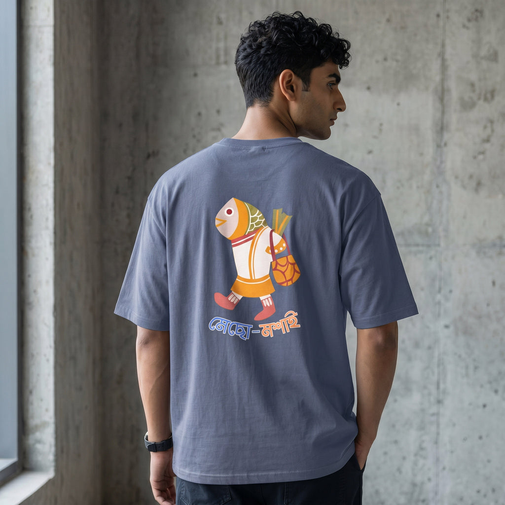 A man wearing a slate blue-gray t-shirt with a colorful cartoon fish graphic on the back, standing in front of a concrete wall.