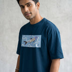A man wearing a navy blue t-shirt with a rectangular graphic print of a stylized fish on a light blue background, standing against a textured gray wall.
