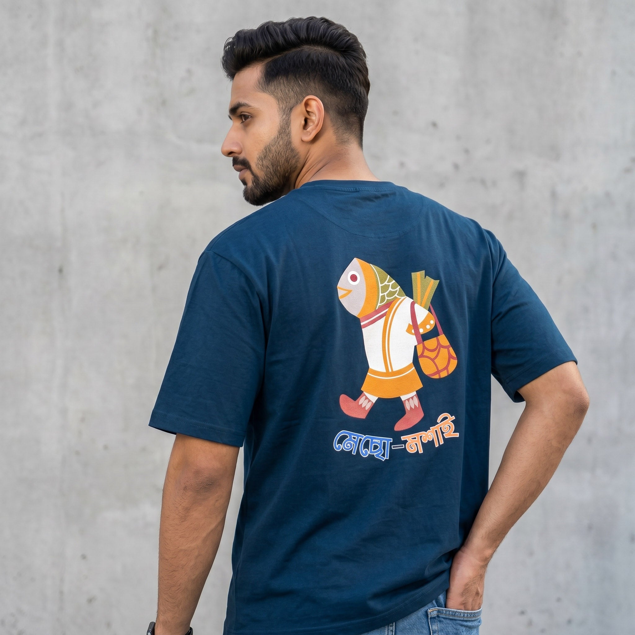 A man wearing a navy blue t-shirt, viewed from the back. The shirt features a colorful cartoon illustration of a fish character carrying a bag, with Bengali text printed below.