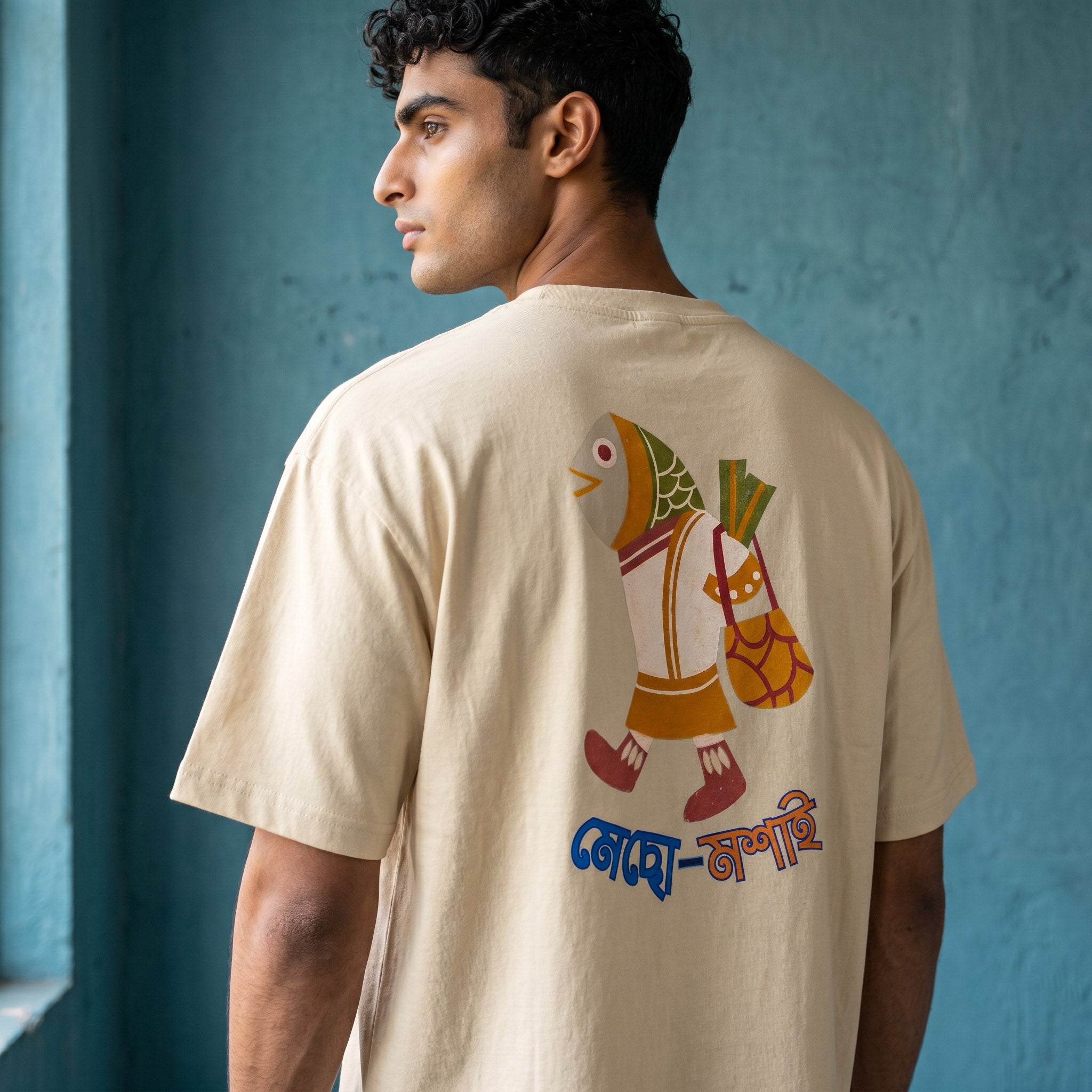 A man wearing a beige t-shirt with a colorful fish graphic and Bengali text on the back, standing against a textured blue wall.