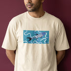 A man wearing a cream-colored t-shirt with a rectangular graphic print of a stylized blue and purple fish swimming in wavy white lines on a teal background, set against a dark burgundy wall.
