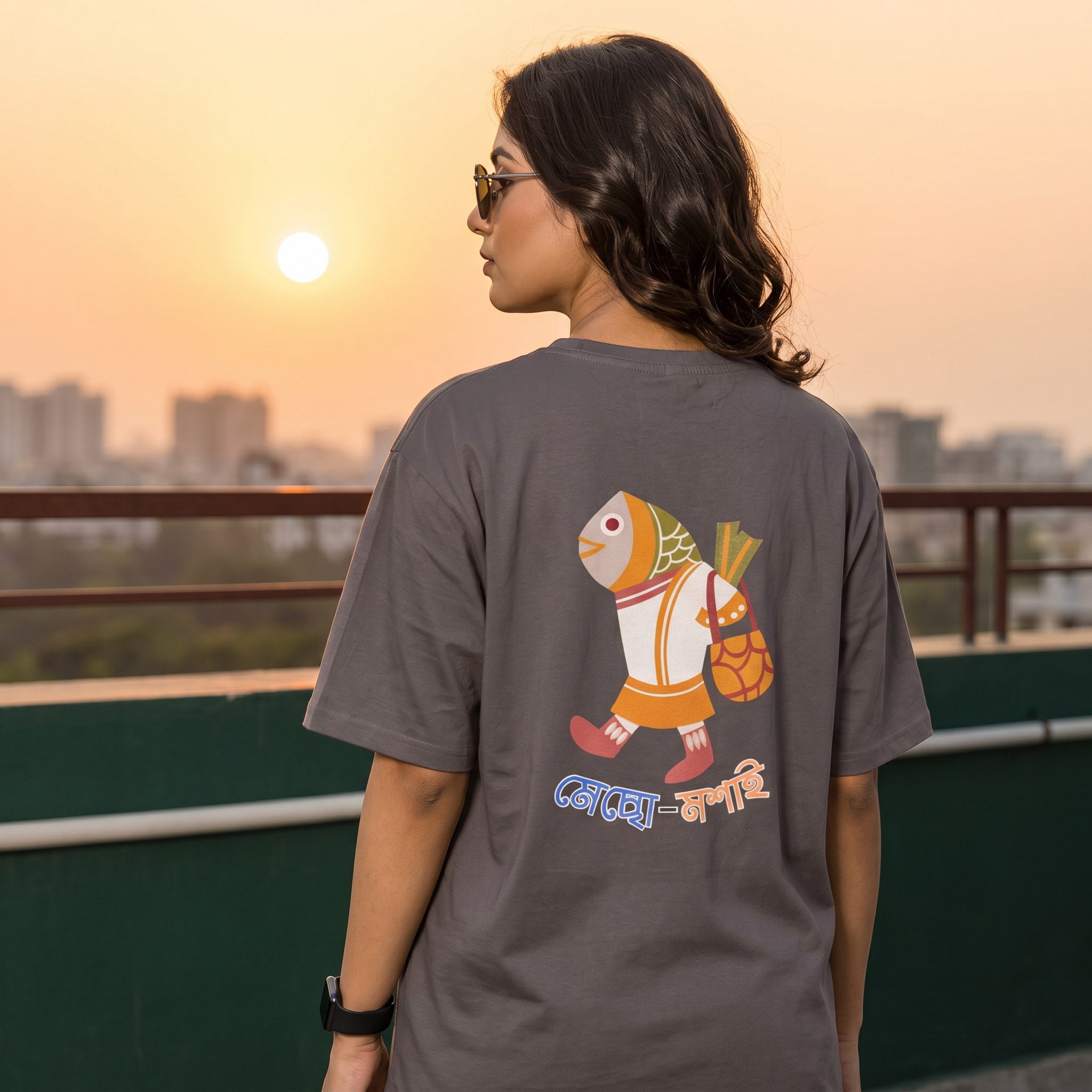 A woman wearing a dark gray t-shirt with a colorful cartoon fish graphic on the back, standing on a balcony at sunset.