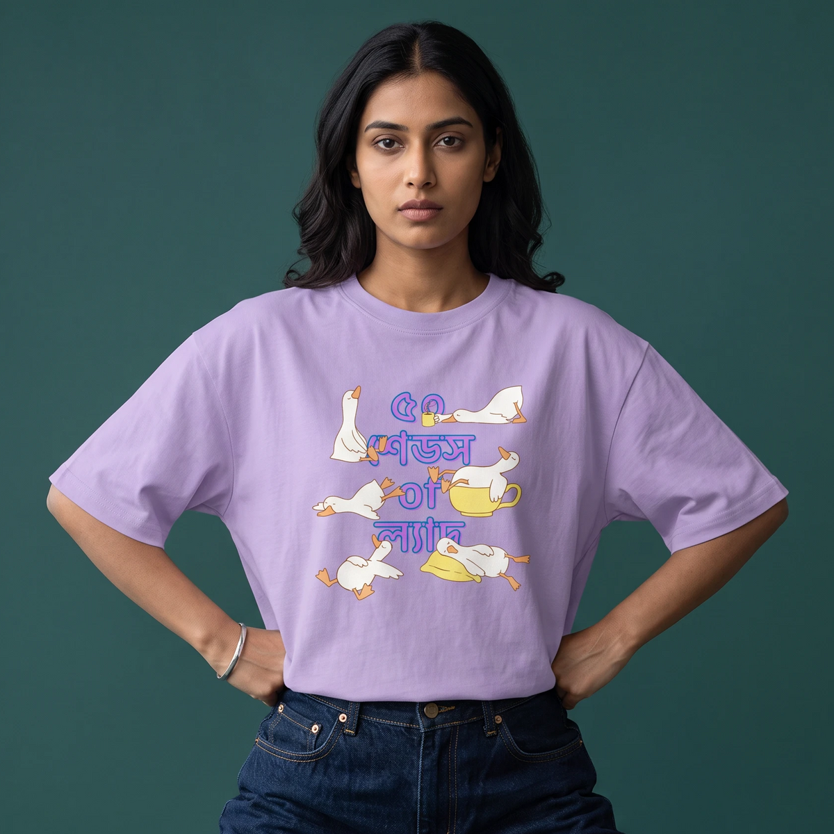 A woman modeling a light purple t-shirt with a cartoon graphic of geese and a teapot in Bengali script, paired with dark blue jeans against a dark green background.