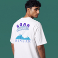 A man wearing a white t-shirt, shown from the back, featuring a graphic print of a blue parachute and mountains with the text 'SOAR BINSAR' in purple and blue, set against a solid dark green background.