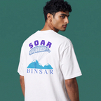 A man wearing a white t-shirt, shown from the back, featuring a graphic print of a blue parachute and mountains with the text 'SOAR BINSAR' in purple and blue, set against a solid dark green background.