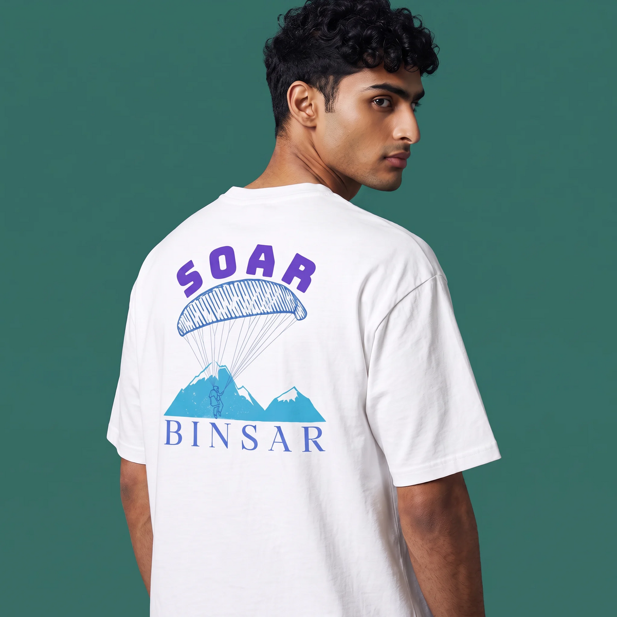 A man wearing a white t-shirt, shown from the back, featuring a graphic print of a blue parachute and mountains with the text 'SOAR BINSAR' in purple and blue, set against a solid dark green background.