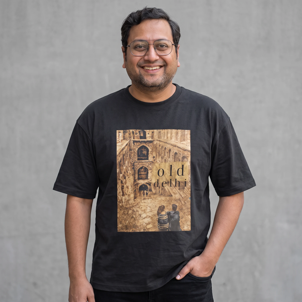 A smiling man wearing glasses and a black graphic t-shirt with a sepia-toned architectural print reading 'old delhi' on the front, standing against a textured gray wall.