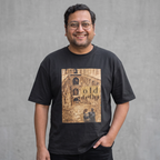 A smiling man wearing glasses and a black graphic t-shirt with a sepia-toned architectural print reading 'old delhi' on the front, standing against a textured gray wall.