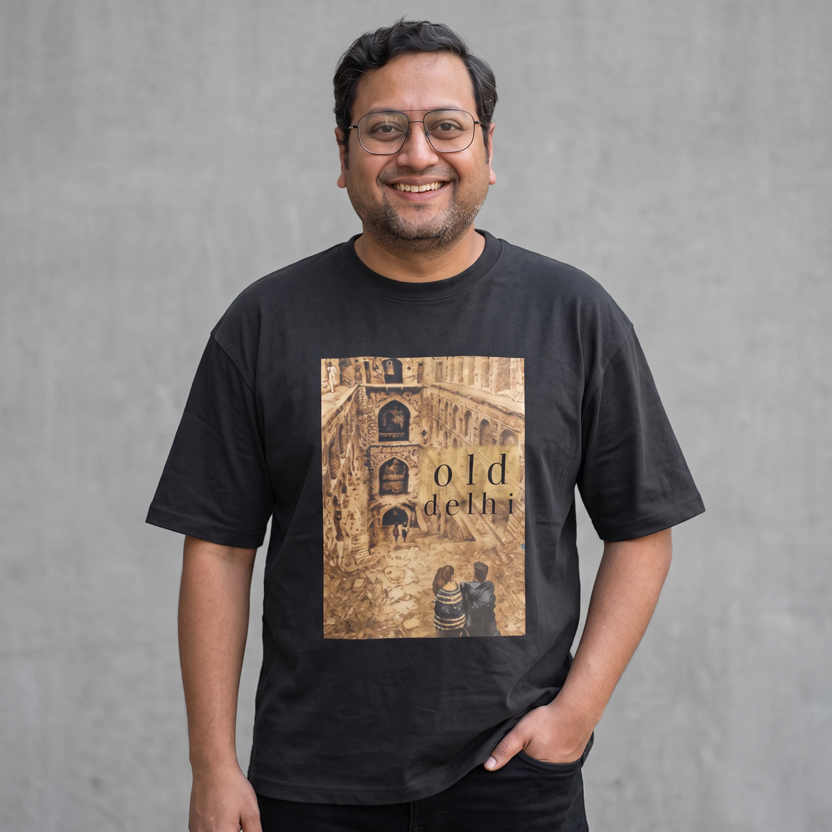 A smiling man wearing glasses and a black graphic t-shirt with a sepia-toned architectural print reading 'old delhi' on the front, standing against a textured gray wall.