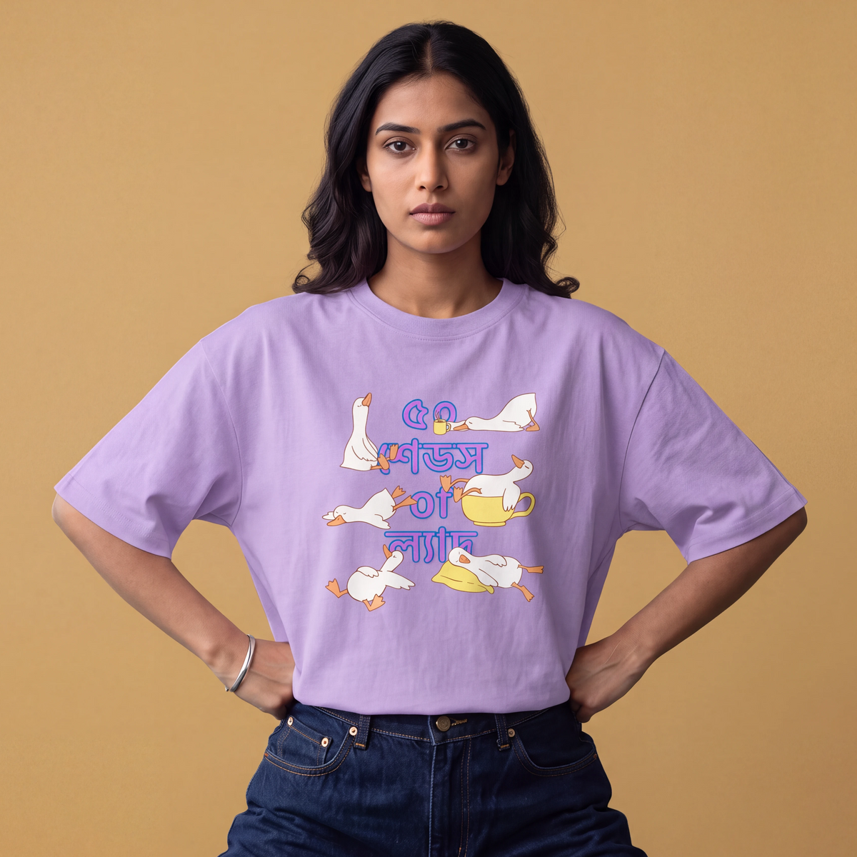 A woman modeling a light purple graphic t-shirt with a cartoon geese and teapot design, paired with dark blue jeans, set against a solid warm tan background.
