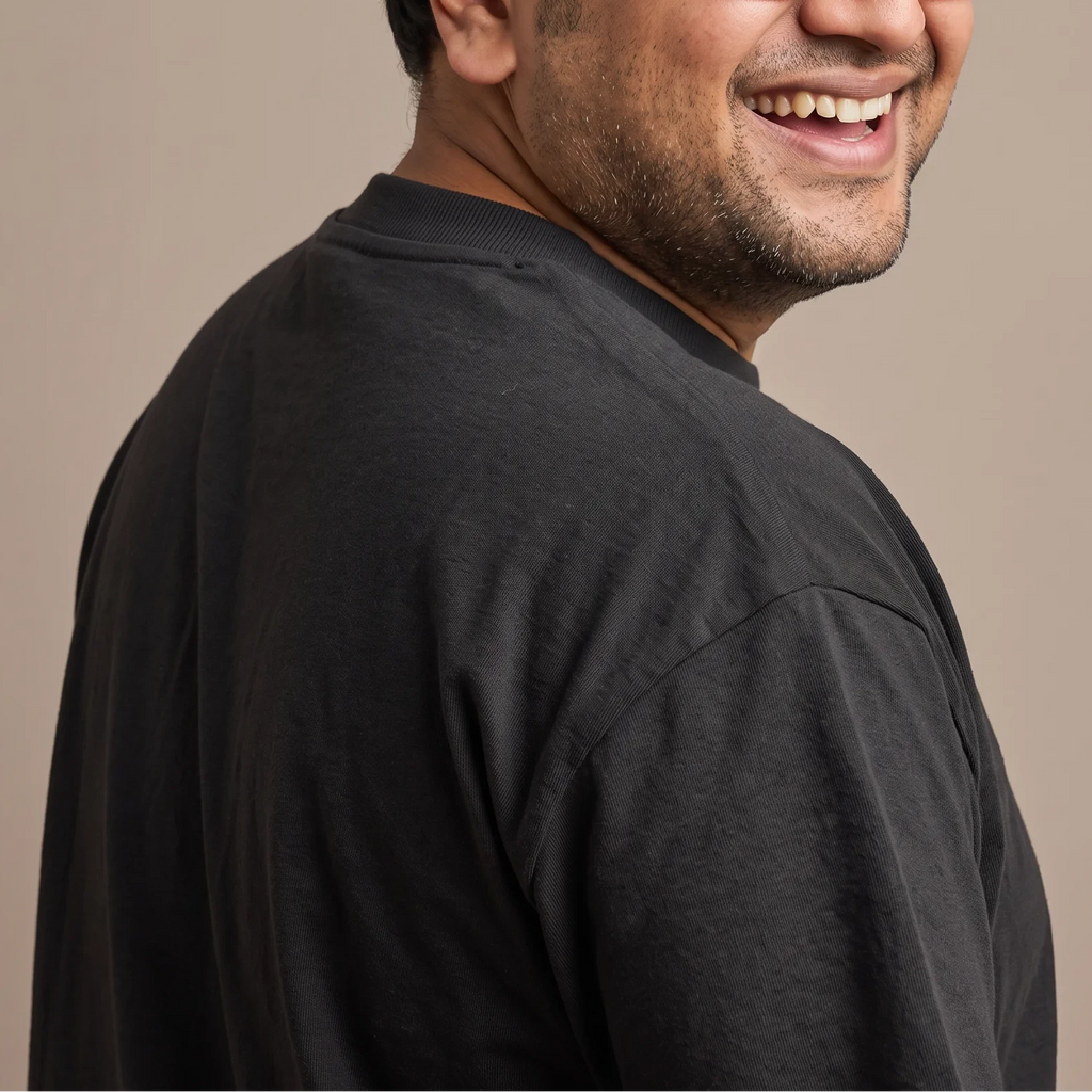 A smiling man with glasses and a short beard, shown in a close-up side profile wearing a dark charcoal gray t-shirt against a solid beige background.