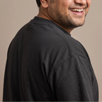 A smiling man with glasses and a short beard, shown in a close-up side profile wearing a dark charcoal gray t-shirt against a solid beige background.